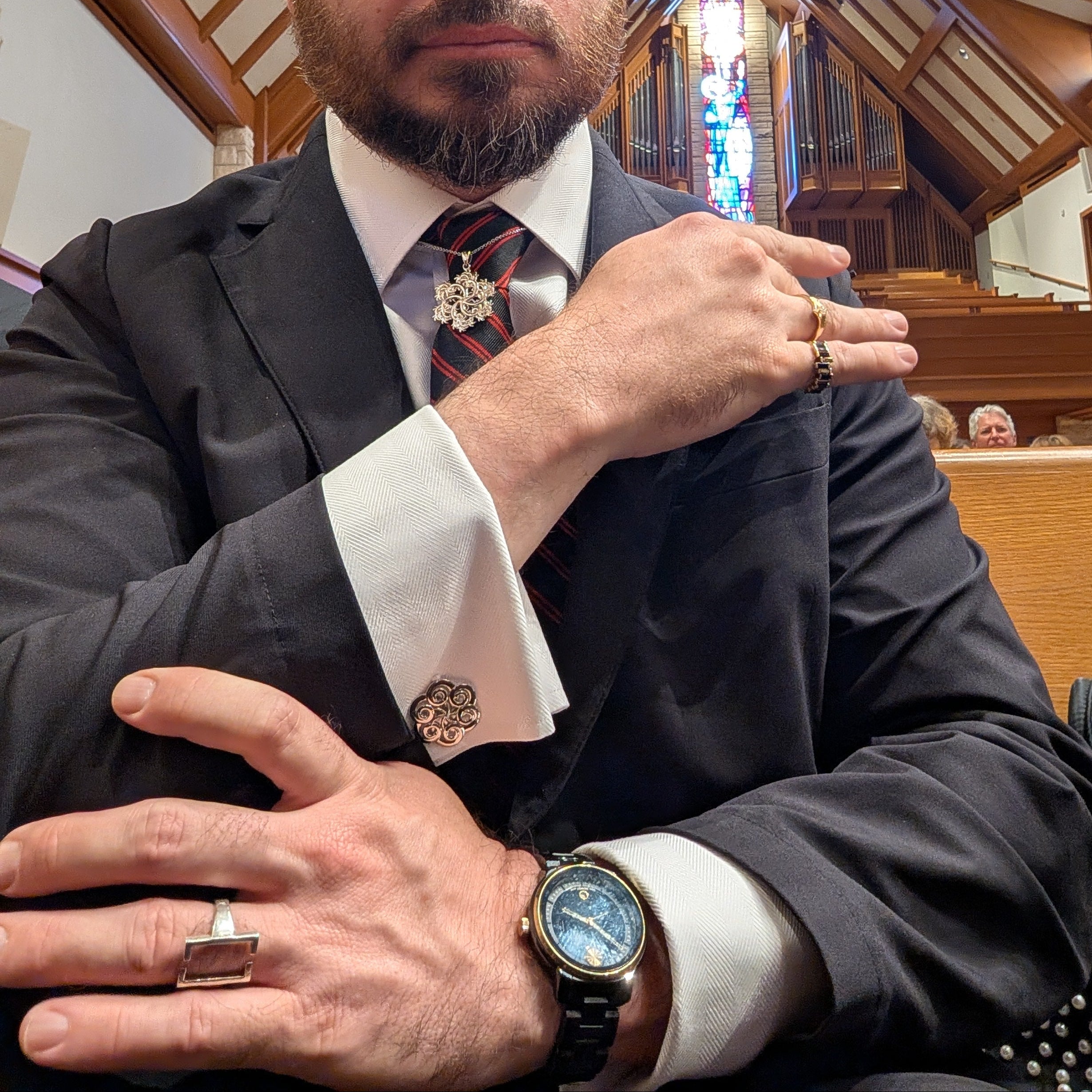 A man in a dark suit, white dress shirt, and striped tie sits in a church pew, gesturing with his hands. The background shows a wooden ceiling and stained glass windows.
