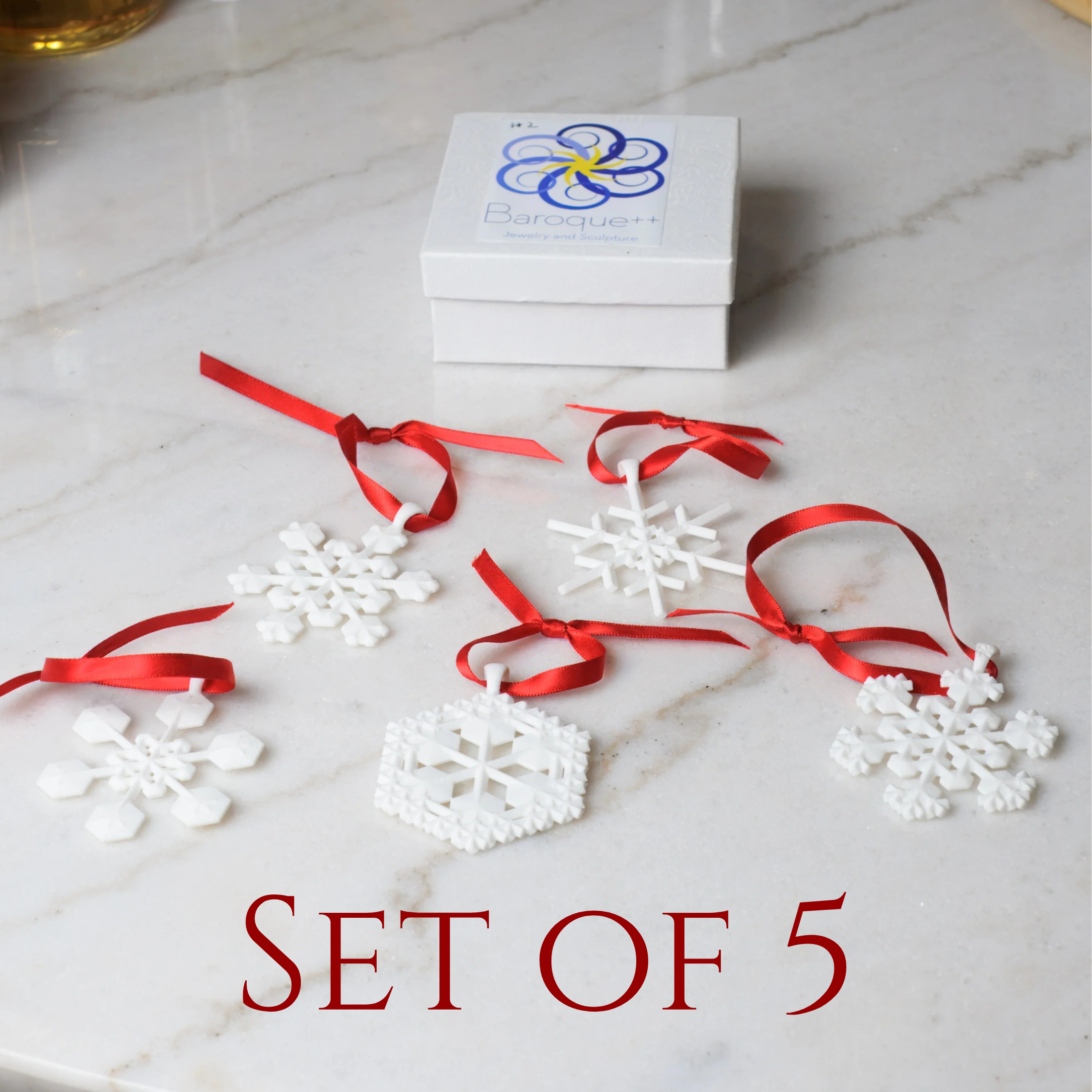 Five white snowflakes with red ribbons on a white marble table. with a white box with logo. Text at bottom says "Set of 5"