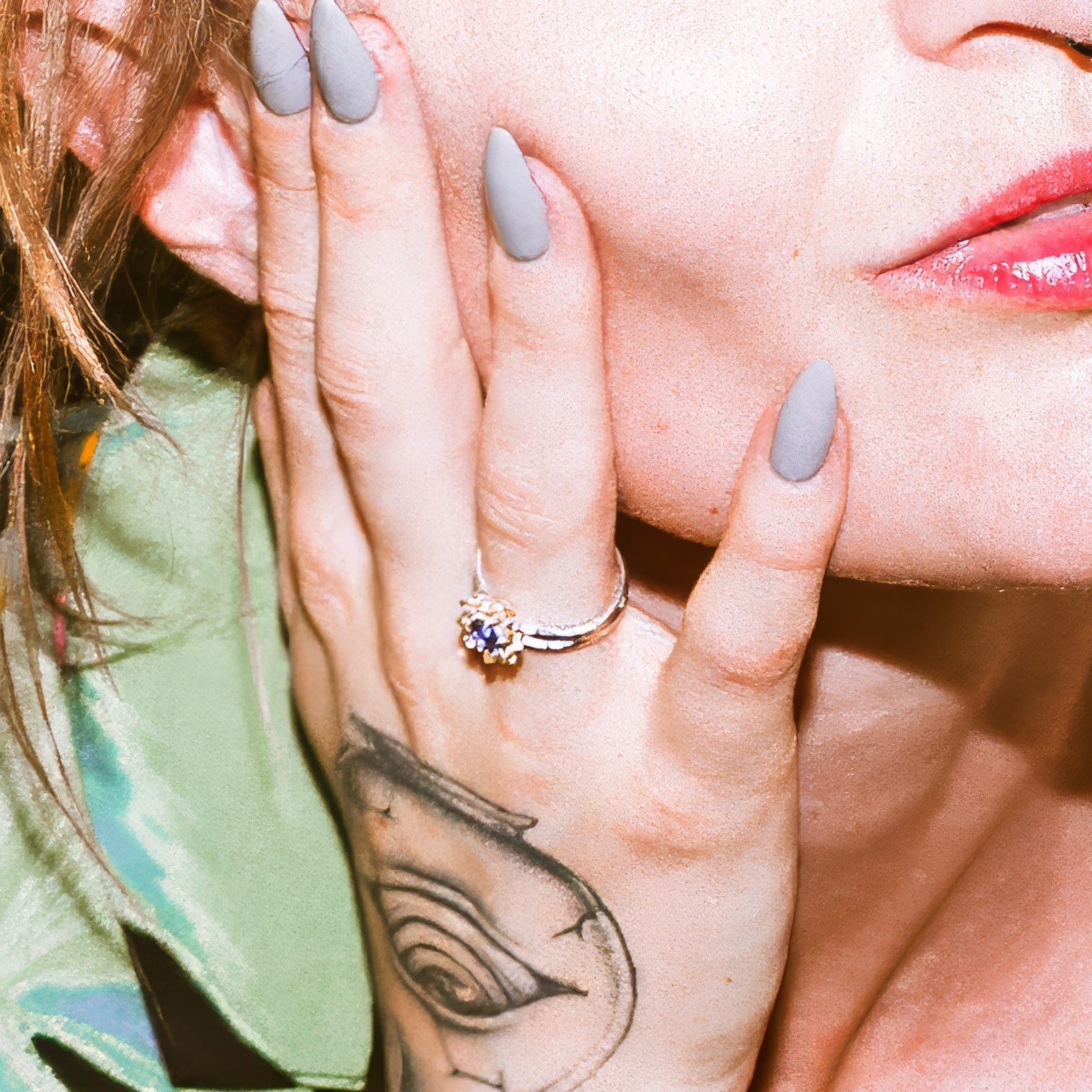 a gold ring with a blue sapphire on a woman's hand touching her cheek. Woman's hand has a tattoo of an eye.