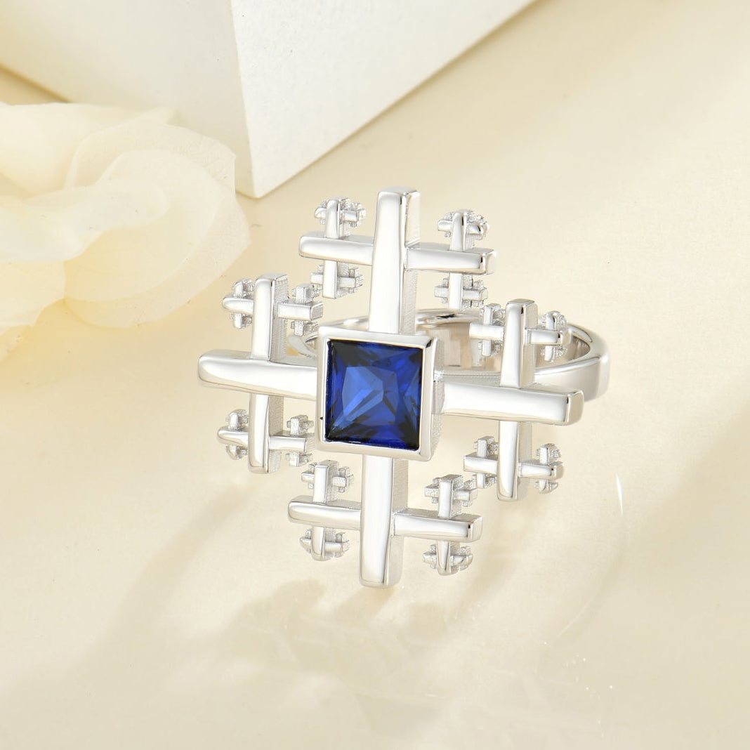 A silver-toned cross-shaped Crux ring with a square blue gemstone center, displayed on a cream-colored surface with soft white decorative flowers and white display blocks in the background.