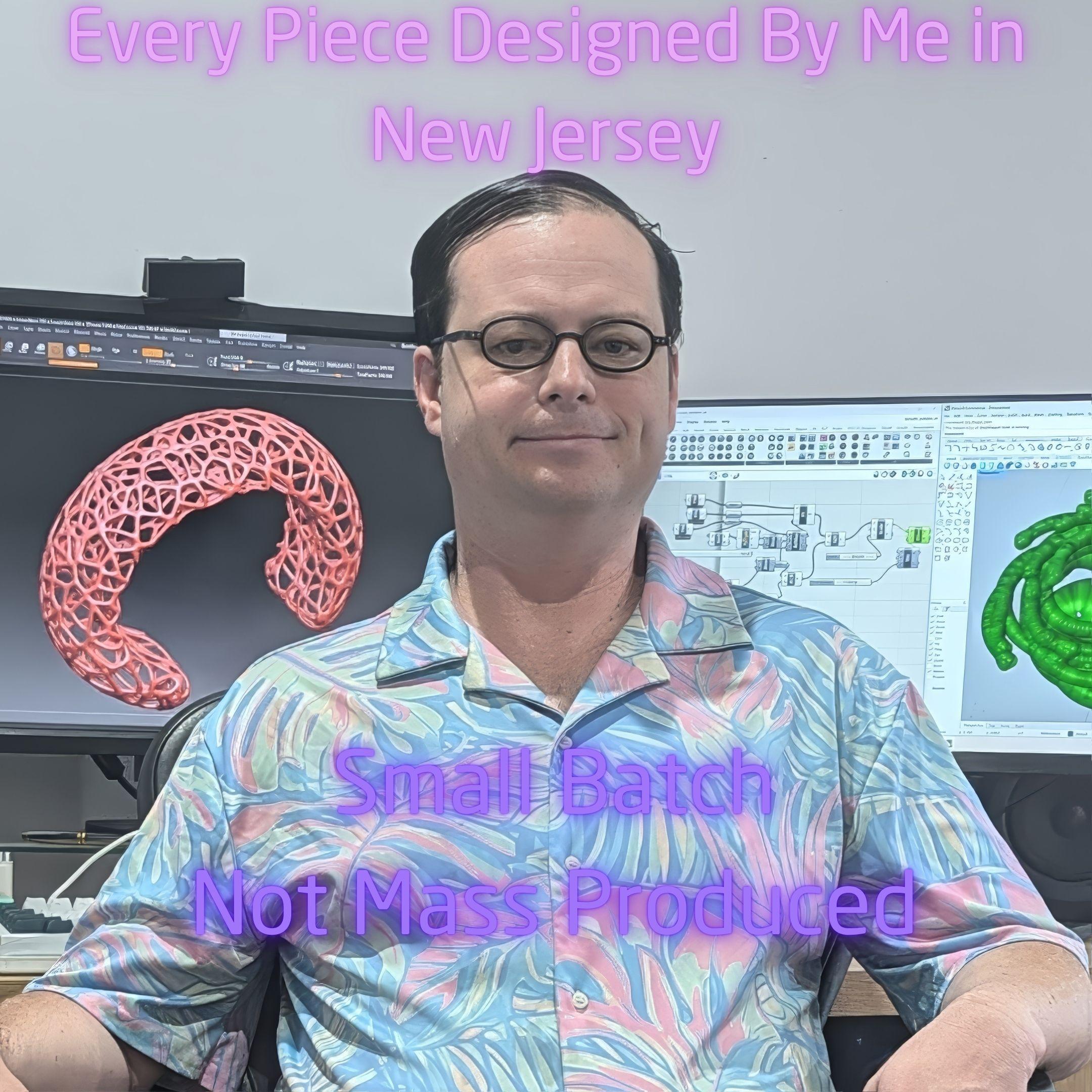 A picture of a man sitting at a desk. Behind him on two monitors are a web-like bracelet on the left, and on the right a visual computer program and a green twist design. The text says "Every piece designed by me in New Jersey. Small Batch Not mass produced.