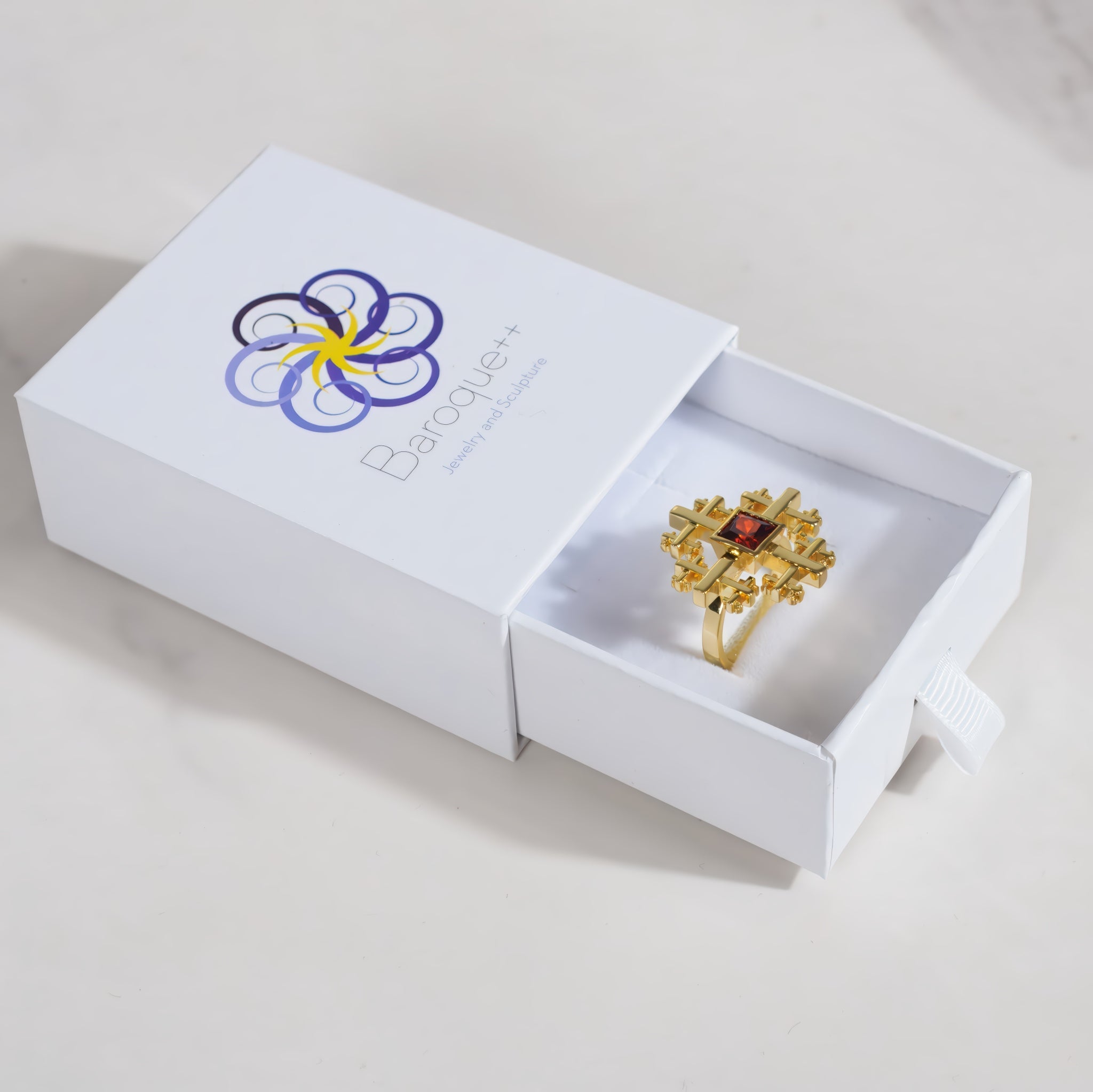A gold Crux cross cocktail style ring with a central red gemstone displayed inside an open white jewelry box branded Baroque jewelry.