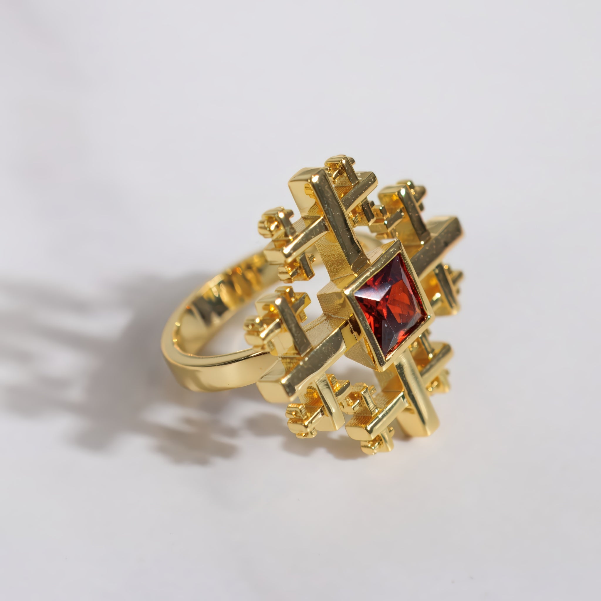 A gold ring with a complex, geometric cross-like design surrounding a central square red gemstone, photographed on a light gray background.