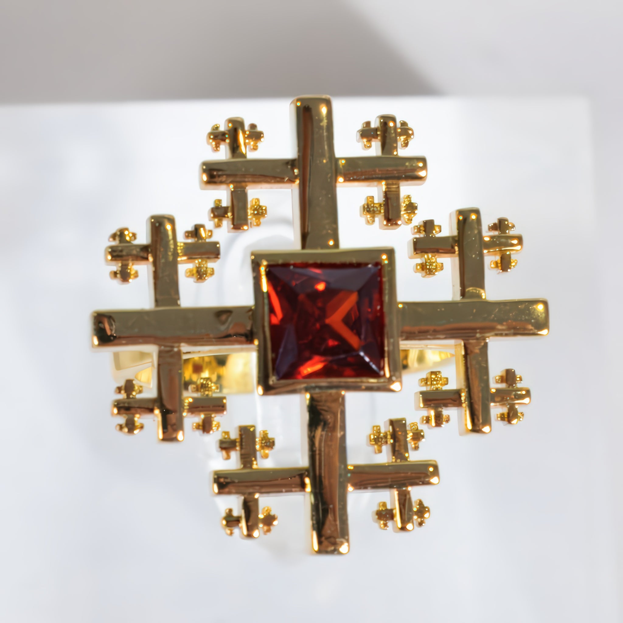 Crux Ring A gold geometric ring featuring a central square-cut dark red gemstone surrounded by an intricate, symmetrical cross-like pattern of smaller gold crosses, displayed on a clean white background.