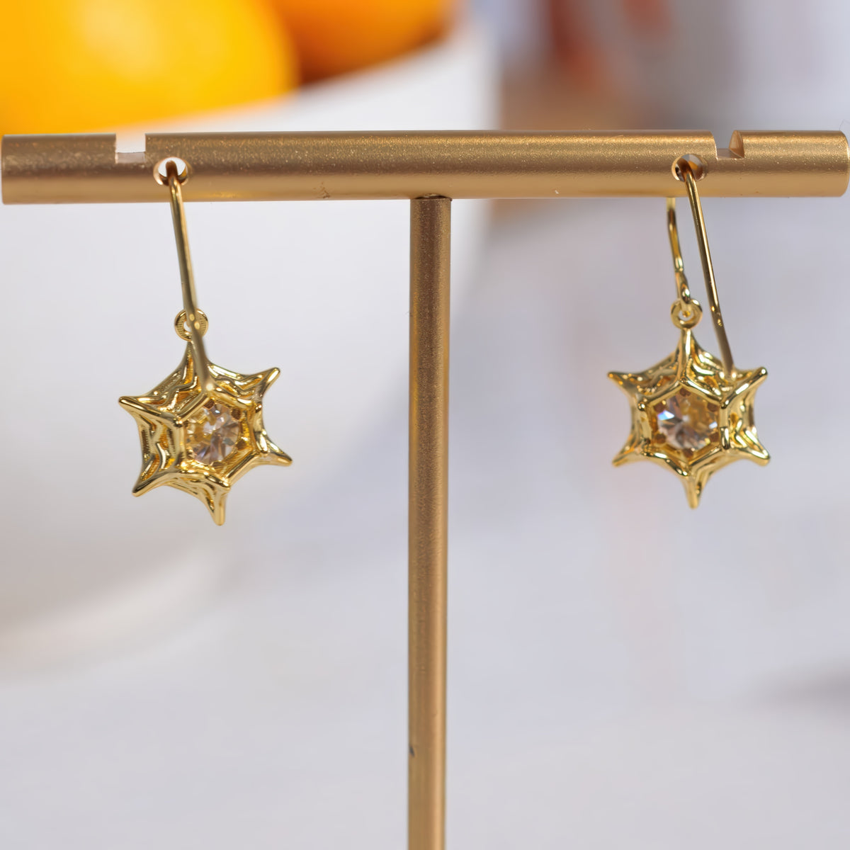 A pair of gold star-shaped dangle earrings with clear faceted center stones, displayed on a gold T-stand against a softly blurred white and orange background.