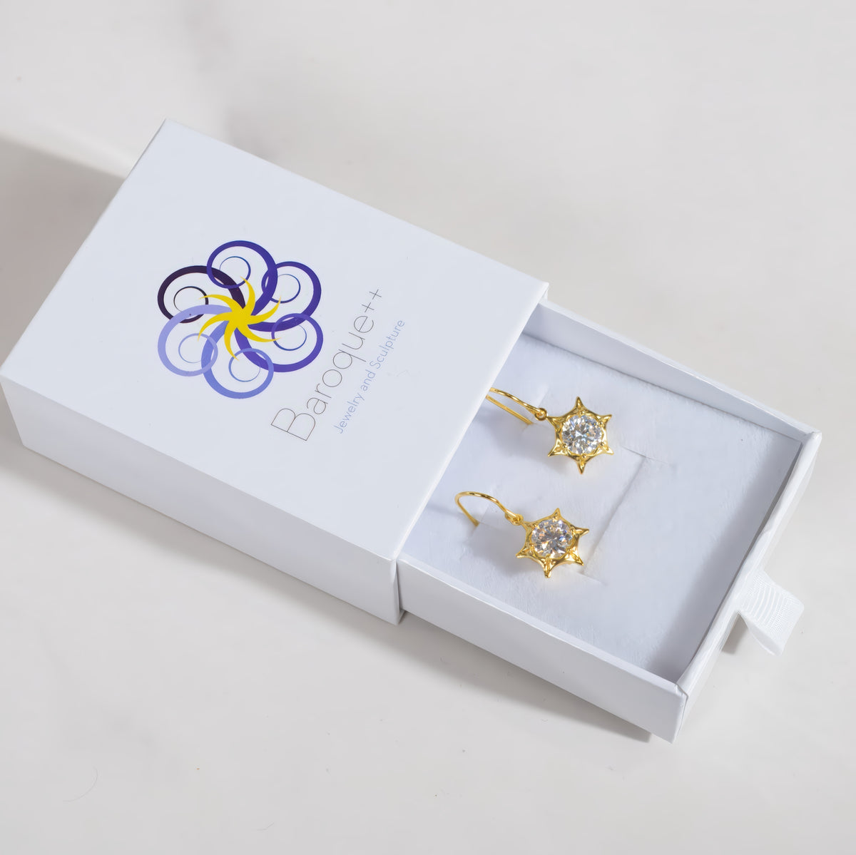 A pair of gold star-shaped dangle earrings with clear stones, presented in a white jewelry box with the brand name Baroque++ visible on the lid.