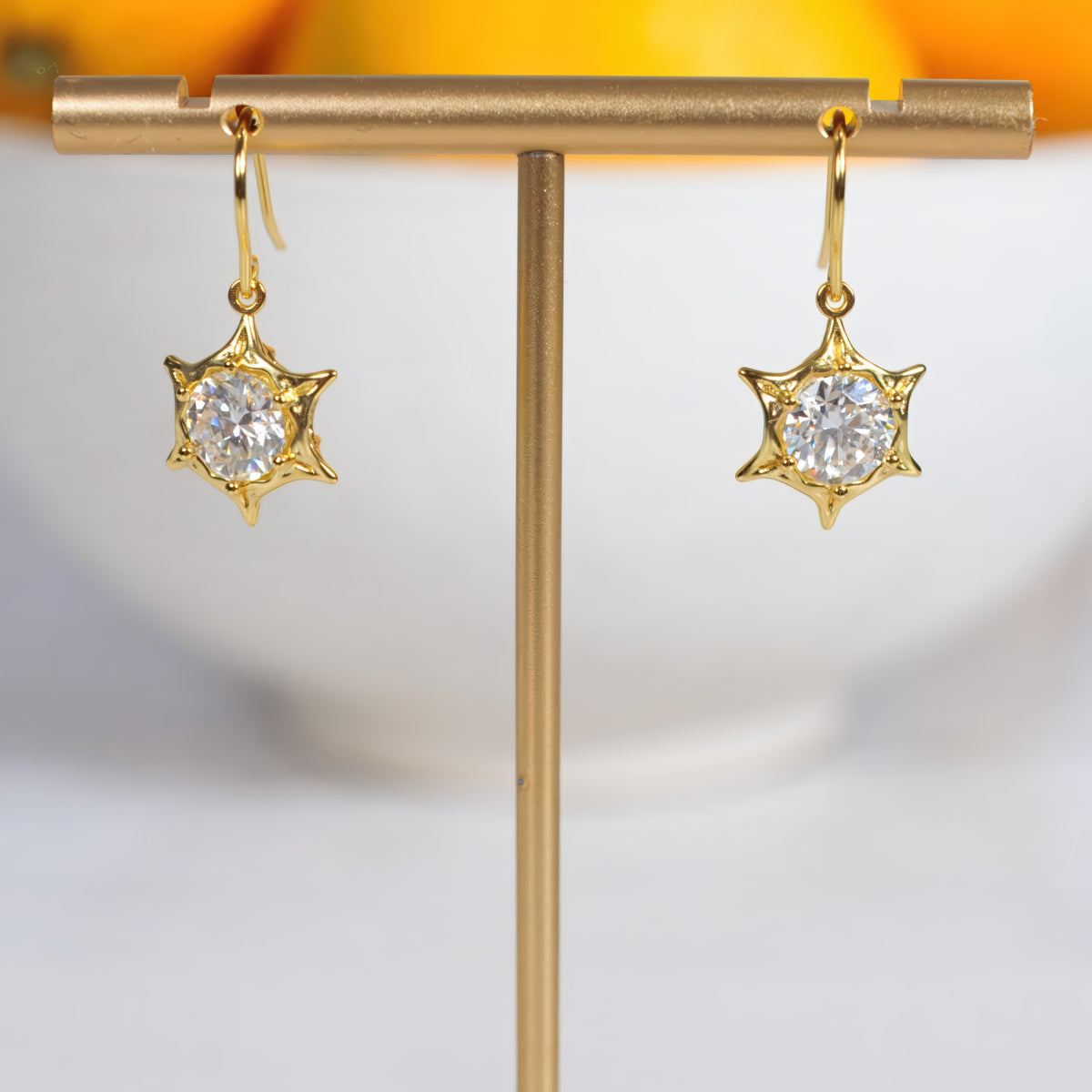 A pair of gold starburst dangle earrings with round clear gemstones, displayed on a gold T-stand against a soft white and orange blurred background.