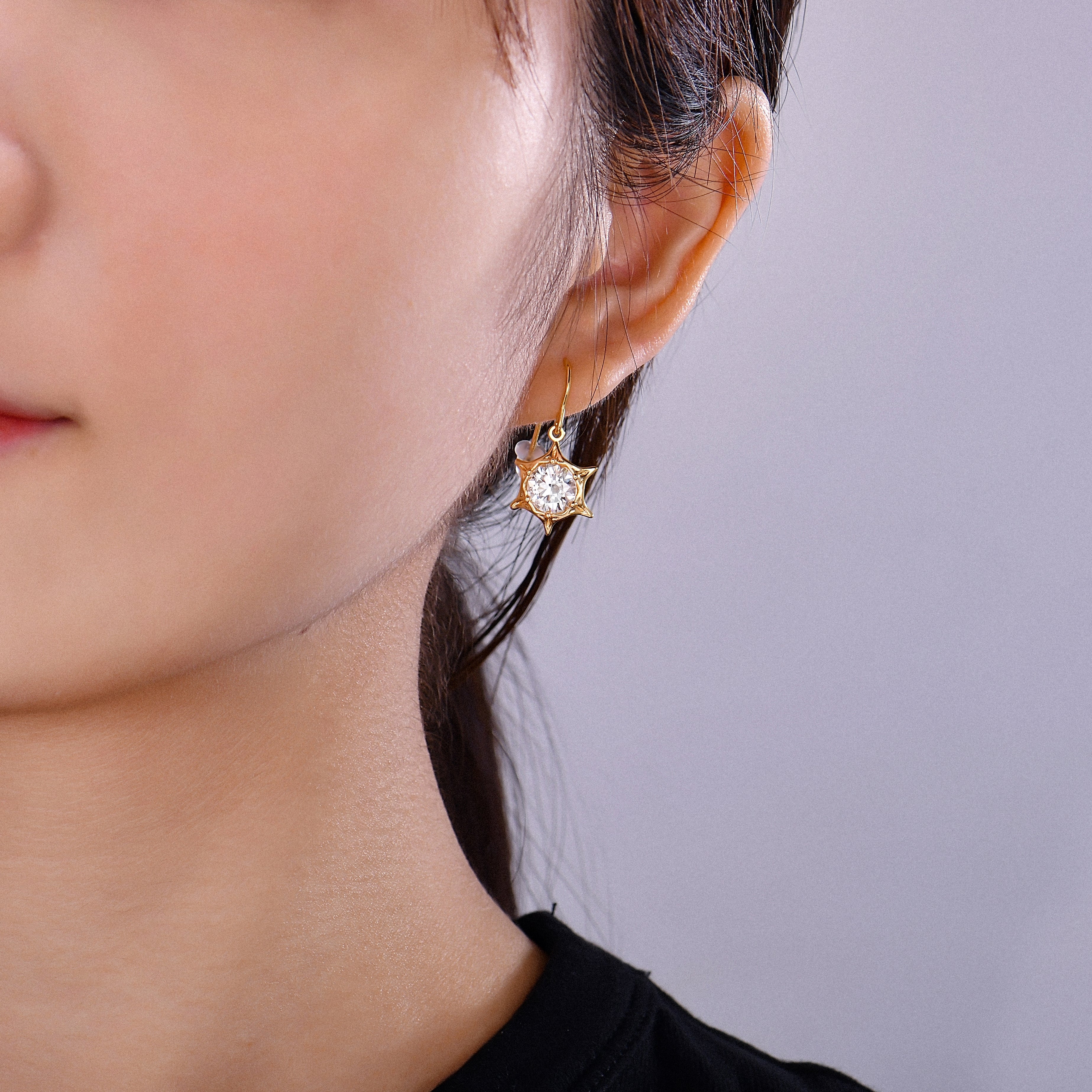 A close-up of a woman's ear wearing a gold cat-shaped drop earring with a clear faceted gemstone, against a plain light gray background.