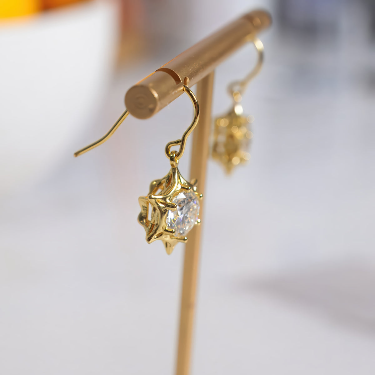 Gold-toned dangle earrings with clear faceted gemstones in starburst-like settings, displayed on a gold jewelry stand against a softly blurred light background.