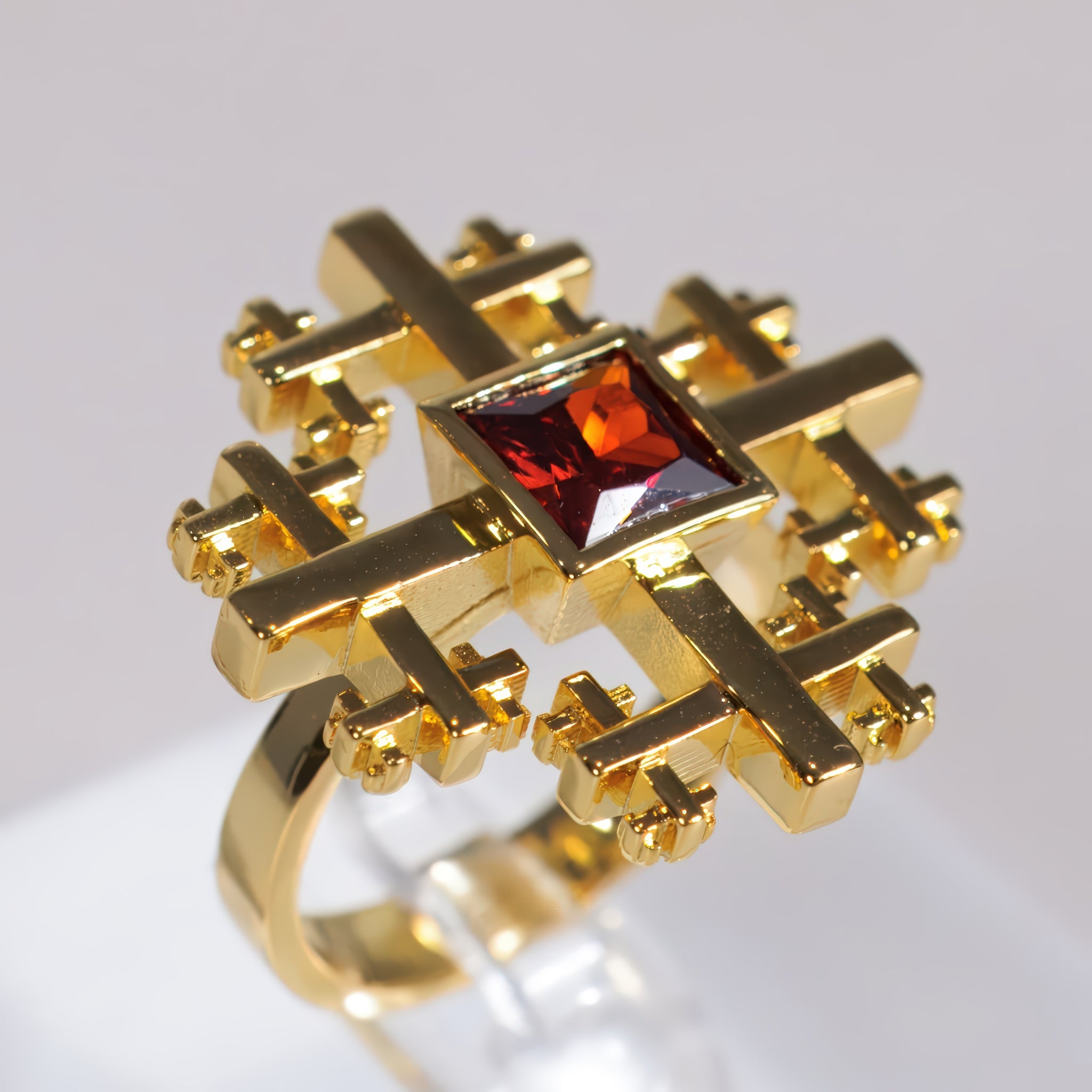 A gold ring Crux Ring with a geometric, cross-like openwork design and a square-cut red gemstone in the center, displayed on a clear stand against a light gray background.