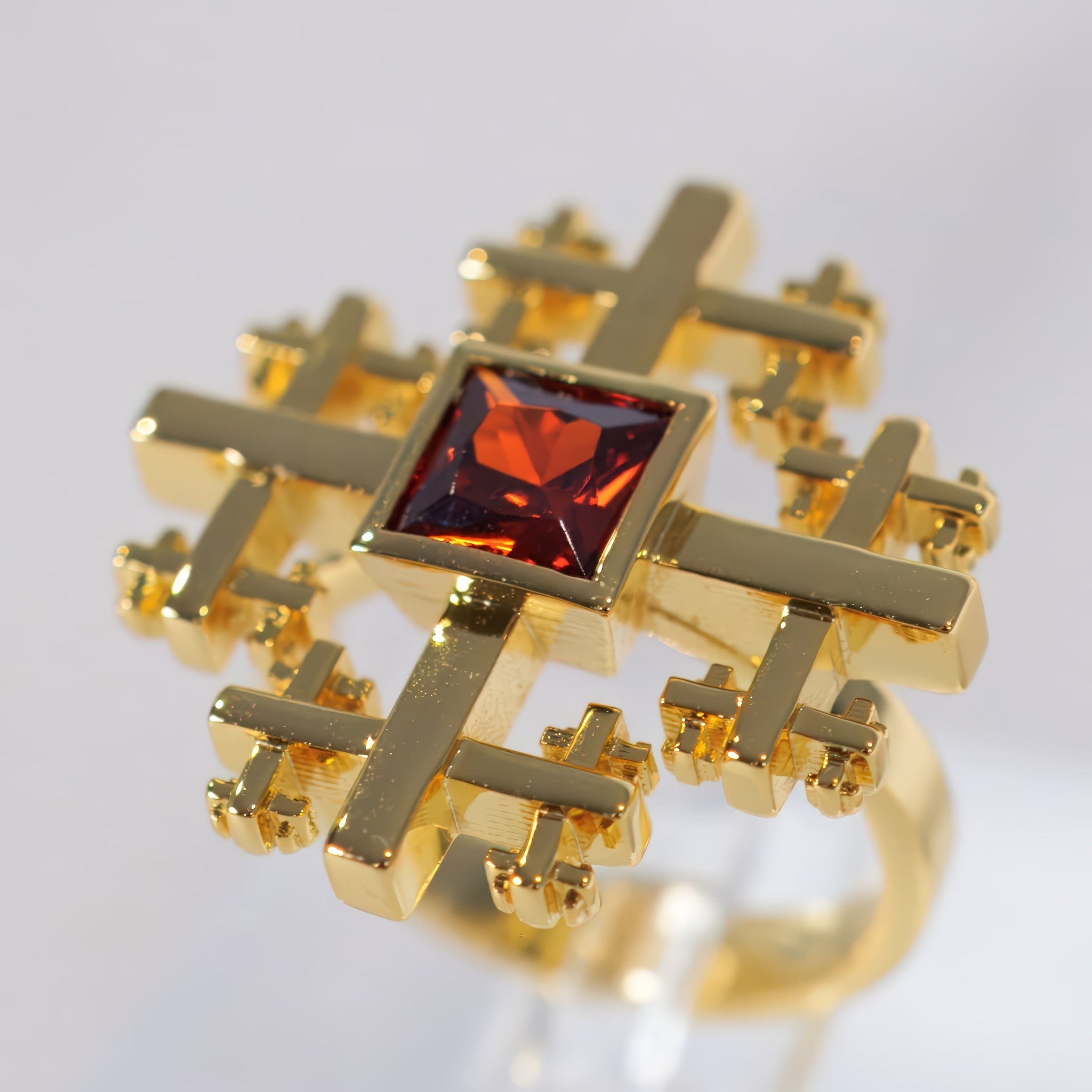 A gold ring Crux Ring with an intricate, geometric cross-like design surrounding a square-cut dark red gemstone, shown on a light gray background.