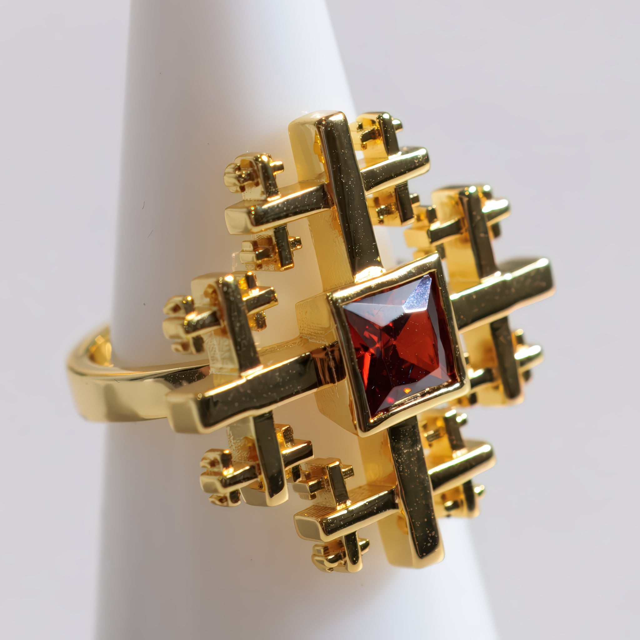 A gold ring Crux Ring with a complex geometric cross-like design and a central square red gemstone, displayed on a white cone-shaped ring holder against a light gray background.