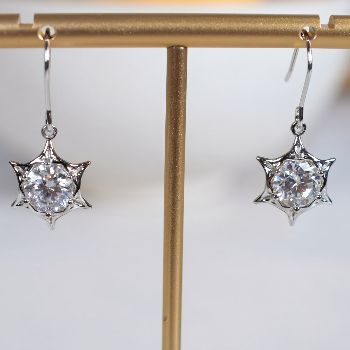 A pair of silver-toned dangle earrings with starburst prong settings holding clear round gemstones, displayed on a gold jewelry stand against a light gray background.