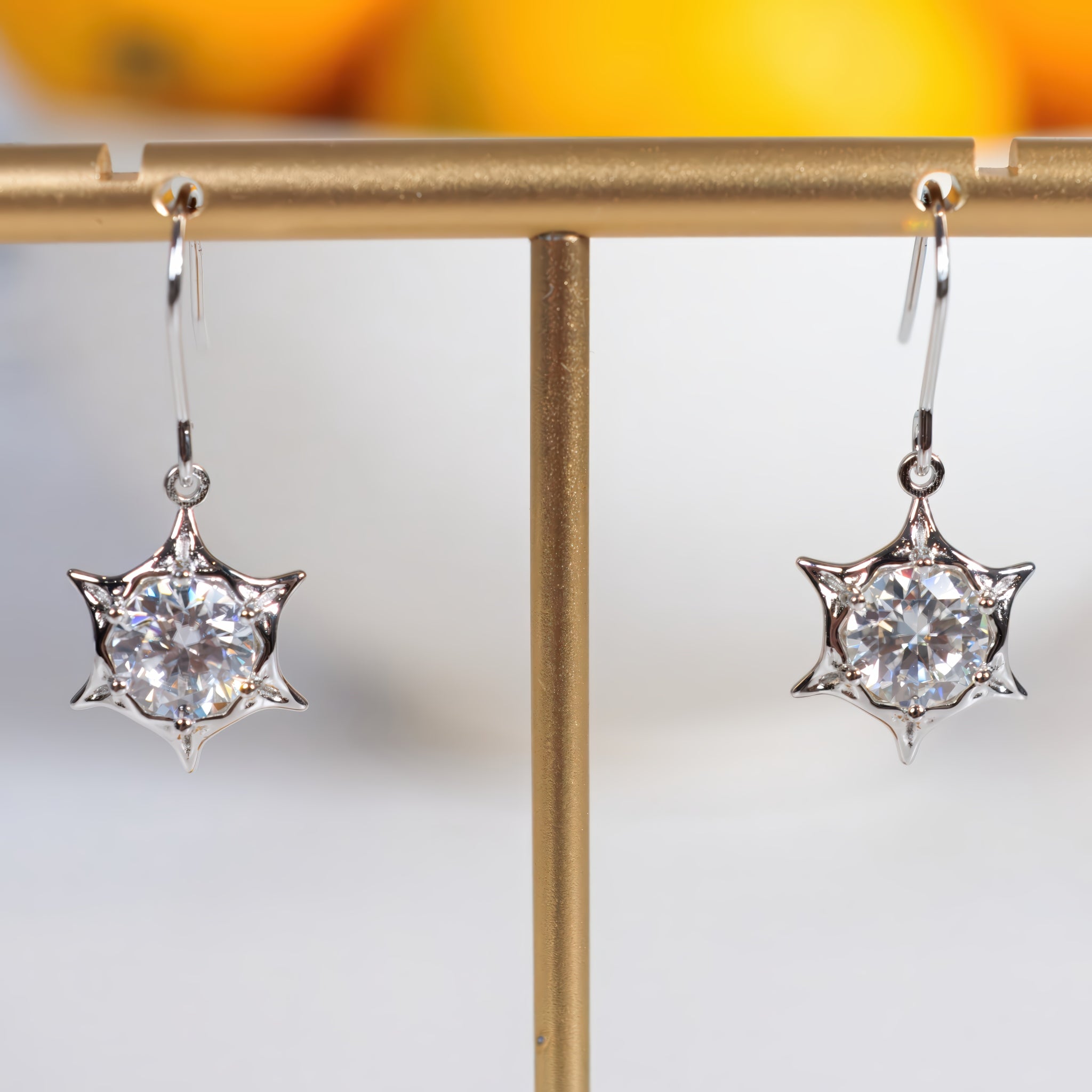 A pair of silver-toned dangle earrings with round clear gemstones in a starburst setting, displayed on a gold jewelry stand against a softly blurred background.