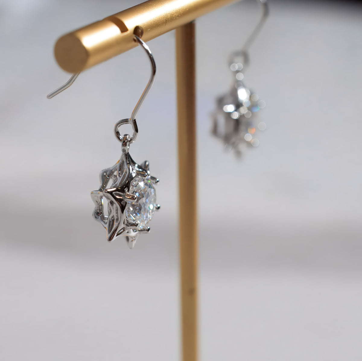 A close-up of a silver-toned dangle earring with a clear faceted gemstone in a starburst-like setting, hanging from a gold display stand against a soft light gray background.