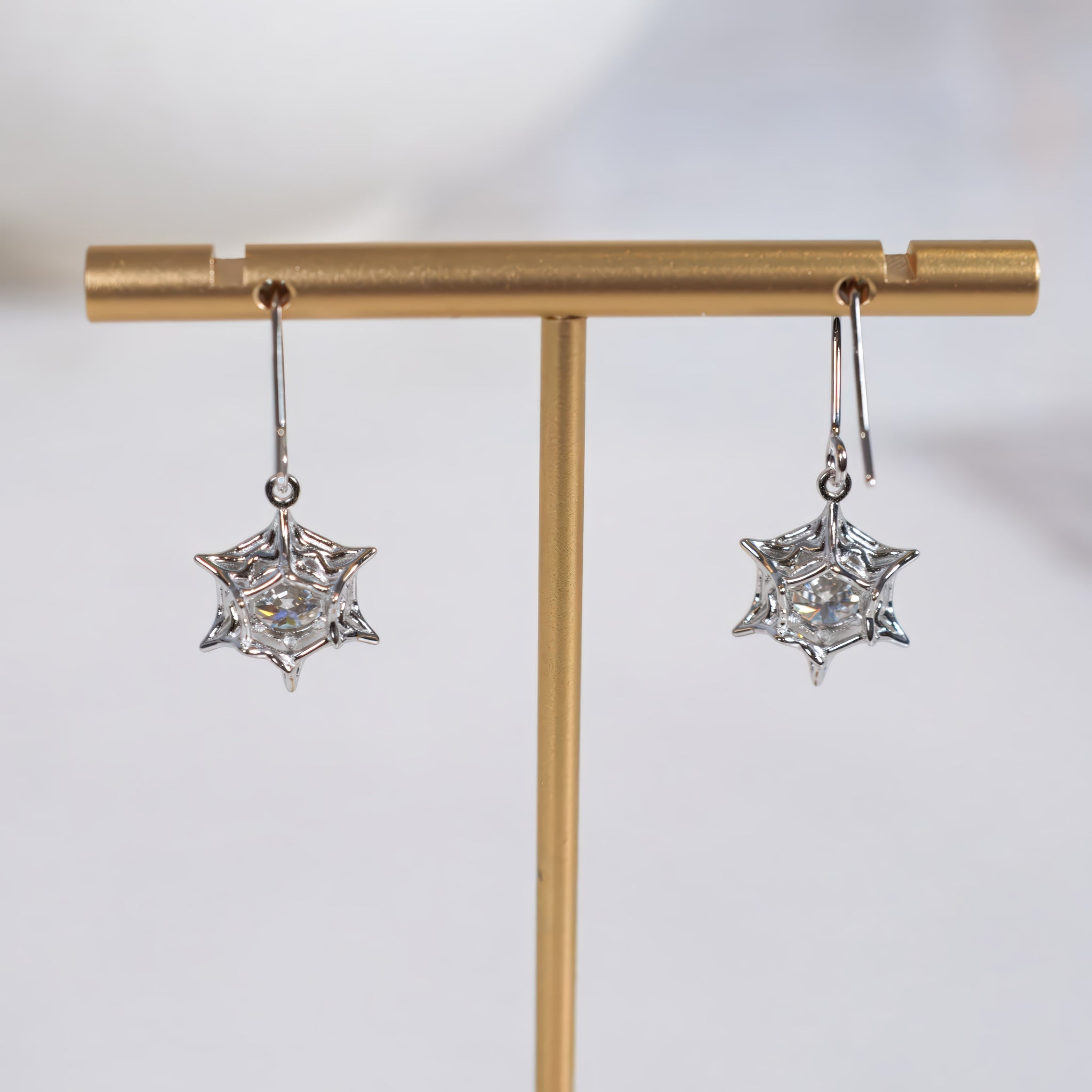 A pair of silver star-shaped dangle earrings with clear faceted gemstones, hanging from a gold T-stand against a light gray background.
