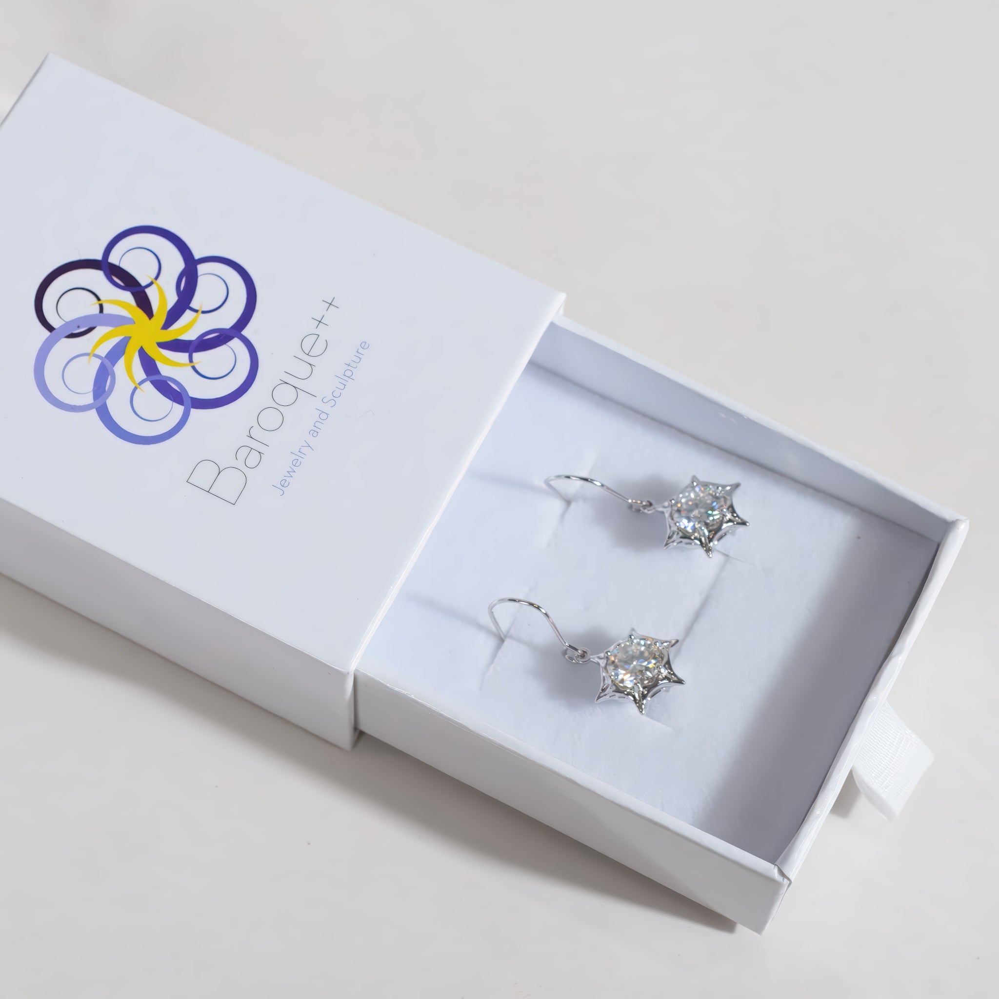 A pair of silver star-shaped drop earrings with clear crystals, presented in a white jewelry box with the Baroque++ logo.