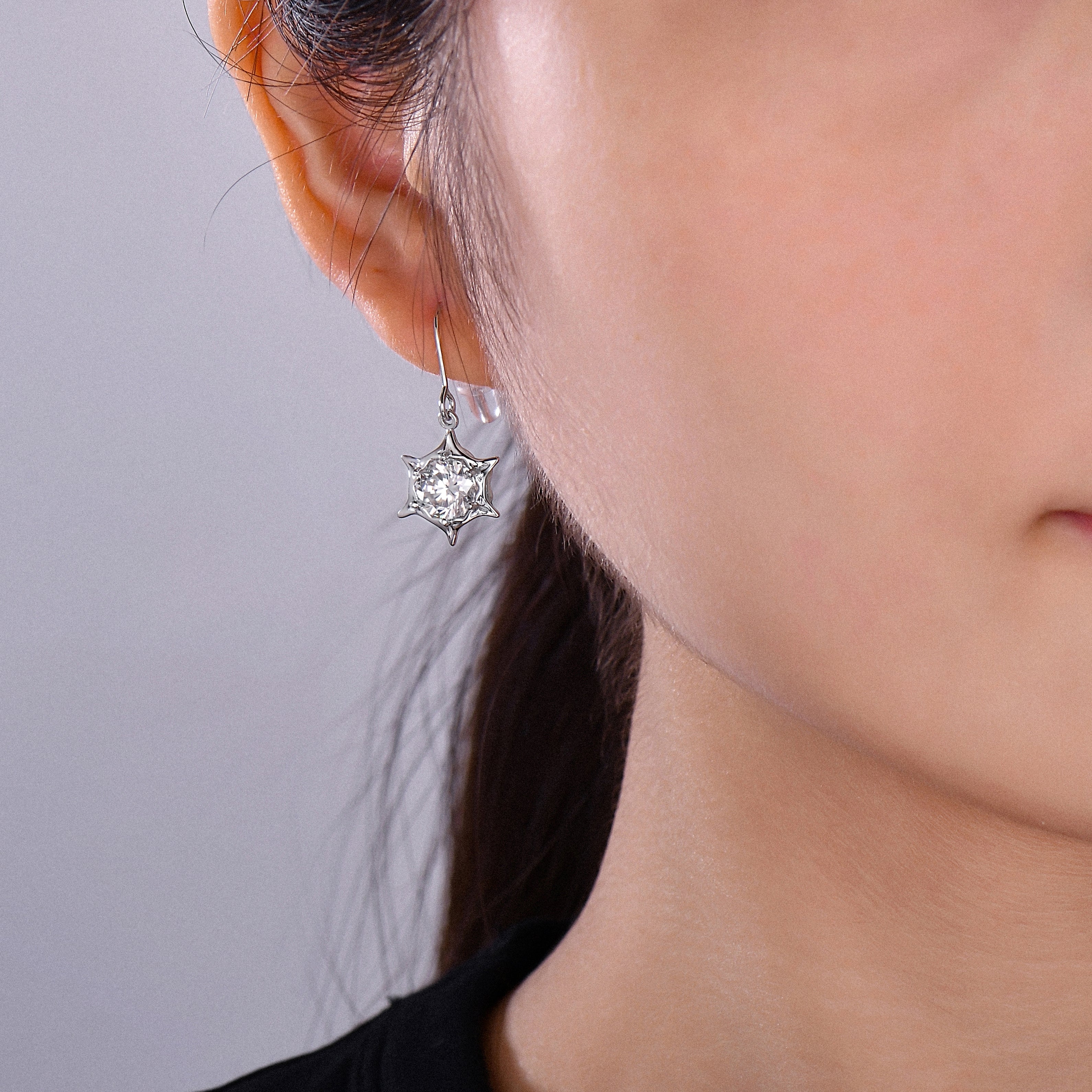 A close-up of a woman's ear wearing a silver-toned dangle earring with a clear faceted star-shaped gemstone.