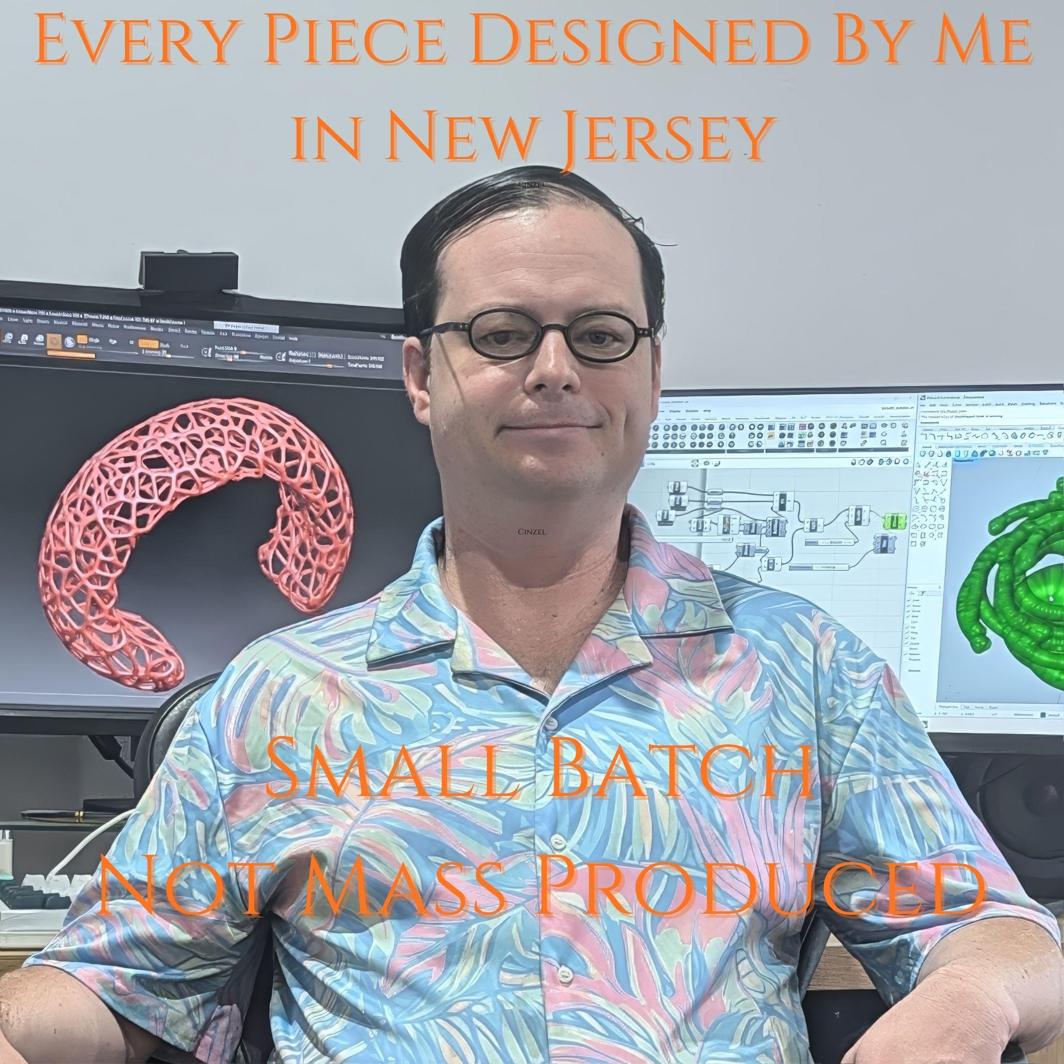 A picture of a man with glasses at a desk. Behind him on the left monitor is an image of a web-like bracelet. On the right monitor is a visual programming interface and a green twisting geometry. The text says "Every Piece Designed by Me in New Jersey. Small Batch Not Mass Produced."
