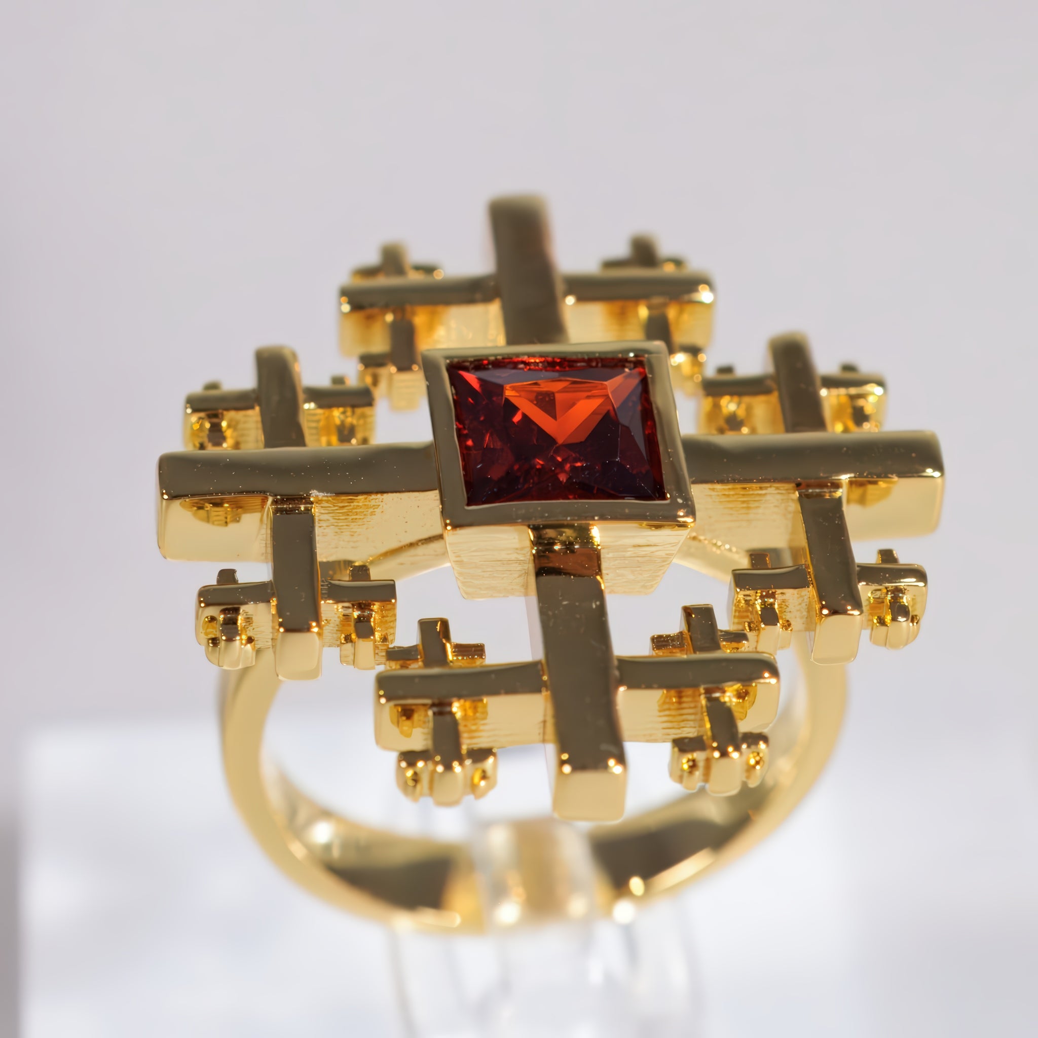 A gold ring Crux Ring with a geometric cross-like design and a central square-cut red gemstone, displayed on a clear stand against a light gray background.
