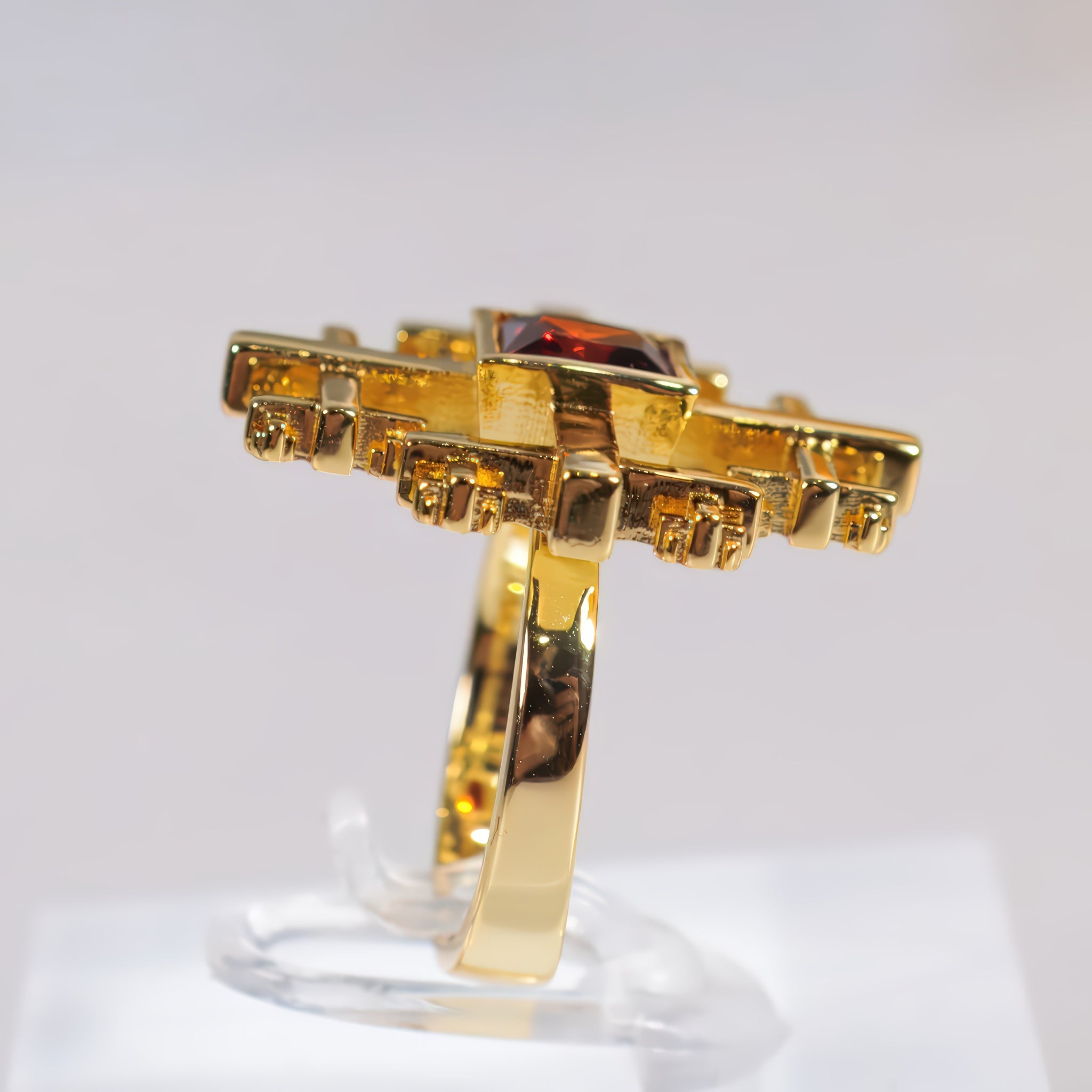 A gold ring Crux Ring with a square dark red gemstone, displayed on a clear stand against a light gray background.