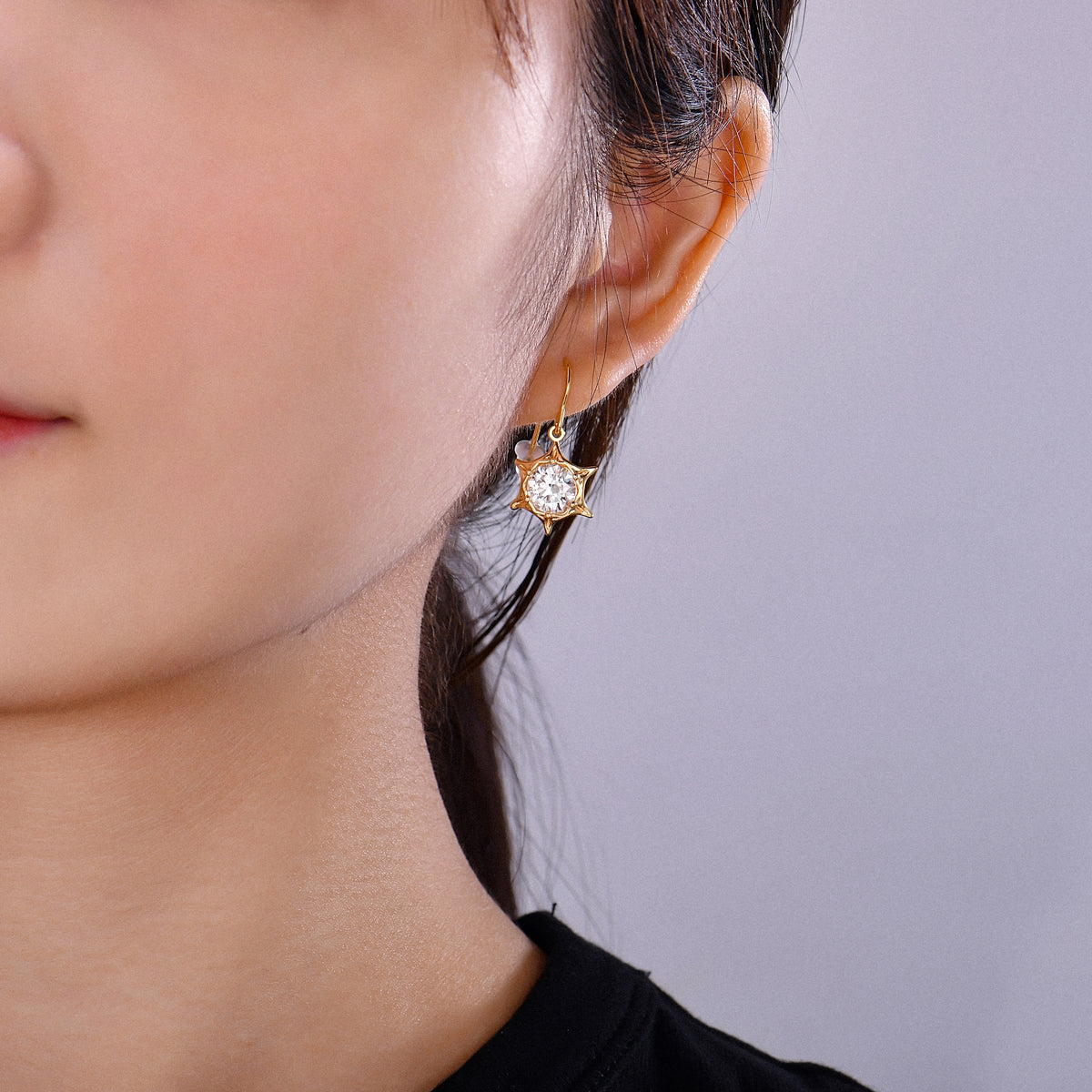 A close-up of a woman's ear wearing a gold cat-shaped drop earring with a clear faceted gemstone, against a plain light gray background.